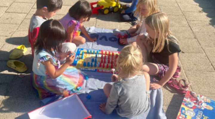 Kleurenfoto van een groepje jonge kinderen die buiten op een zonnige dag op een betegeld pleintje een bordspel spelen.