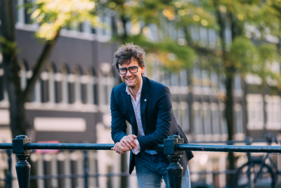 Kleurenportretfoto van Levi van Dam. Hij kijkt lachend de camera in, met zijn onderarmen steunend op een brugleuning. Op de achtergrond zijn gebouwen en bomen te zien.